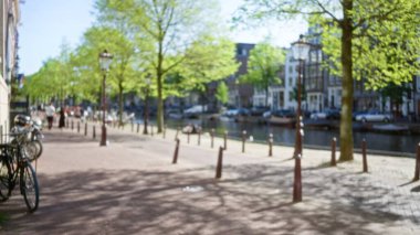 Arka planda bisikletler, ağaçlar ve tarihi mimariyle güneşli bir kanalın bulanık görüntüsü, hareket halindeki insanlarla birlikte tipik bir şehir günü sahnesini yakalıyor..