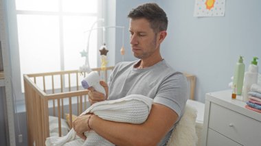 Genç adam bebeği yatak odasında tutuyor, beşiğin yanında termometreyle oturuyor, evin içinde huzurlu bir babalık anı yakalıyor.
