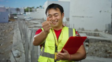 Bir inşaatta kalp hareketi yapan genç bir adam, güvenlik yeleği giyiyor ve dışarıda bir pano tutuyor, bitmemiş bir inşaat alanında kameraya gülümsüyor..