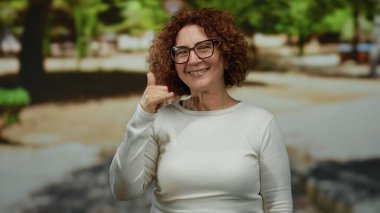 Orta yaşlı İspanyol bir kadın açık hava parkında kameraya bakıp gülümseyen, doğal ışık altında mutlu bir konuşma öneren bir jest yapıyor..