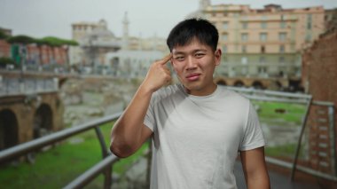 Şaşkın bir hareketi olan genç adam, şehir mimarisi ve yeşilliklerle çevrili hareketli bir caddede duruyor, açık havada şaşkınlığını ifade ediyor..