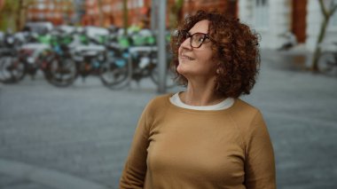 Kalabalık bir caddede arka planda bisikletlerle gülümseyen bir kadın gülümseyen bir ifade ve canlı bir açık hava atmosferiyle sıradan bir şehir manzarası sergiliyor..