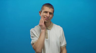 Çenesi ağrıyan bir adam mavi arkaplana karşı yanak tutarken arkasında izole bir duvar olan sıradan bir gömlek giyiyor..