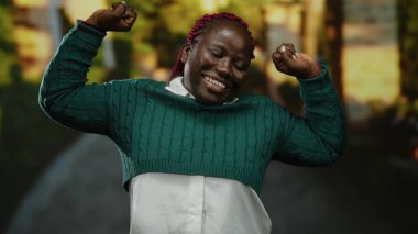 Güneşli bir parkta yeşilliklerle eğlenen, rahat bir kazak giyen, neşeli ifadesi ve açık havada yaptığı hareketlerle mutluluğu ve kendine güveni gösteren bir kadın..