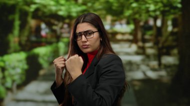 Yumruklarını sıkmış gözlüklü, kendine güveni tam bir İspanyol kadın, canlı bir park ortamında duruyor. Etrafı yemyeşil ve güneşli bir patikayla çevrili..