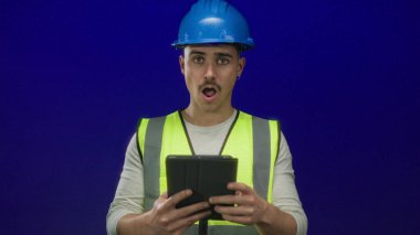 Yansıtıcı yelek ve şapka giyen genç İspanyol adam, izole edilmiş mavi bir arkaplanda bulunan tablete şaşırmış görünüyor. Profesyonel bir ortamda beklenmedik bir keşif anı yakalıyor..