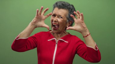 Kırmızı elbiseli, gri saçlı yaşlı bir kadın canlı yeşil bir arka planda pençeli ellerle neşeli bir şekilde kükreyerek enerji ve coşku saçıyor..