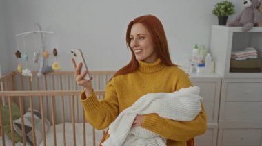 Bebek beşiği ve oyuncak ayıyla sıcak yatak odasında selfie çekerken kucağında bir kadın, modern mobilyalarla huzurlu bir evde anne şefkati gösteriyor..