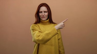 Sarı bir süveteri basit bir kahverengi duvar arka planına doğrultup kafa karışıklığını ifade eden bir kadın, kırmızı saçlarına ve günlük tarzına odaklanmış duygu jestleri sergiliyor.