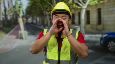 Şehir caddesinde bağıran, güvenlik yeleği ve şapka takan Asyalı genç adam, şehir dışında inşaat sahnesini gösteriyor, hareketli bir ortamda iletişim enerjisi ve nişan taşıyor..