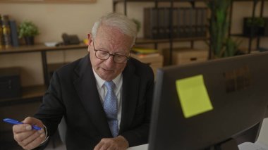 Takım elbiseli kıdemli bir adam, kitap ve bitkilerle çevrili bir ofis ortamında özenle çalışıyor, bilgisayara odaklanıyor ve kalemle notlar yazıyor..