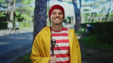 Sarı ceketli ve elinde olta tutan kırmızı bereli genç adam arka planda bulanık ağaçlarla parkta gülümsüyor, boş zamanlarını yakalıyor..