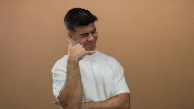 Genç adam rahat bir şekilde gülümsüyor. Yumuşak kahverengi bir arka plana karşı. Çekici ve arkadaş canlısı bir görünüm sergiliyor..