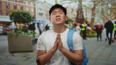 Sırt çantalı genç adam, kalabalık bir şehir caddesinde elleri sımsıkı bağlanmış bir şekilde, sanki şehir dışında dua ediyormuş gibi duruyor..