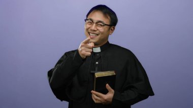 Gözlüklü ve siyah cüppeli Asyalı bir rahip mor bir duvara karşı elinde bir İncil tutuyor ve kameraya güvenle bakıyor..