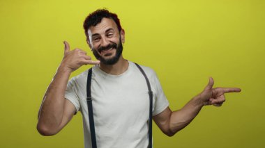 Sakallı bir adam şakacı bir tavırla sarı arka planda beyaz gömlek giyiyor telefon konuşmasını taklit etmek için elleri kullanıyor ve eğlenceli etkileşimli bir anı işaret ediyor.