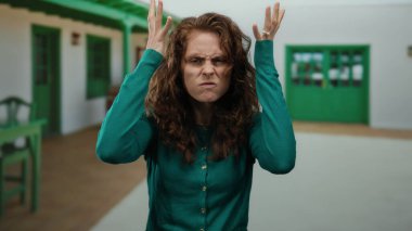 Şehir caddesinde yeşil kazaklı, kıvırcık saçlı bir kadın, açık havada beyaz binanın yanında yeşil aksanlı ve güneşli bir ışıkla tiyatro ifadeleri sergiliyor.