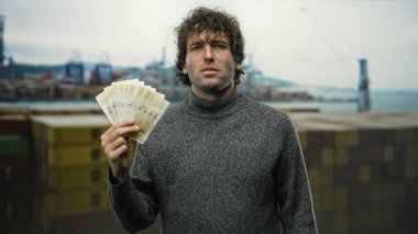 Norveç kronu taşıyan bir adam deniz kıyısında arka planda botları olan, kazak giyen, açık hava düzeni kuran para ve deniz atmosferinin özünü yakalıyor..