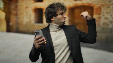 Genç adam, açık hava ortamında elinde akıllı bir telefonla, siyah ceket ve bej balıkçı yaka giyip açık havada azim ve tarz sergileyerek kendinden emin bir jest yapıyor..