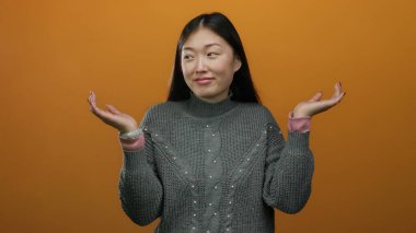 Stüdyo ortamında turuncu arka plan ile izole edilmiş süveter giyen, omuz silken bir kadın belirsizliği ya da sorgulama tutumunu öne sürüyor..