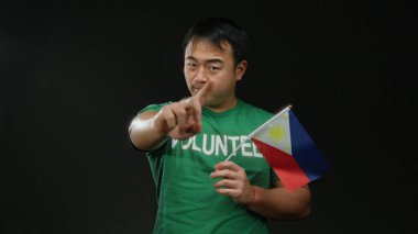 Yeşil gömlekli genç adam siyah arka planda Filipin bayrağı tutuyor. El hareketleri yapıyor. Gönüllülüğü ve ulusal gururu temsil ediyor..