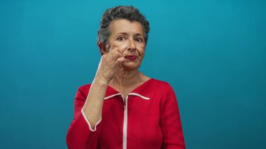 Kırmızı elbiseli yaşlı kadın, ağzını kapatıp mavi arka plana kilitlenmek için jest yapıyor..