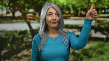 Gri saçlı, mavi kazak giyen son sınıf öğrencisi kadın, açık havada, açık yeşil bir parkta doğal ışık altında sakin bir günün tadını çıkarırken baş parmak işareti yapıyor..