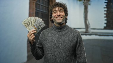 Kentsel bir sokak ortamında Amerikan dolarlarını tutarken gülümseyen İspanyol adam, şehrin arka planında finansal başarı ve mutluluk sergiliyor..