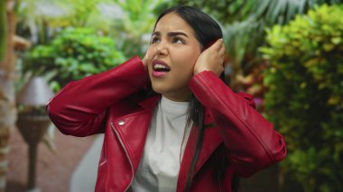 Kırmızı ceketli, elleri kulaklarını tıkayan, yemyeşil parktaki kadın. Etrafı canlı doğa ile çevrili, dışarıdaki duyguları yansıtan biri.