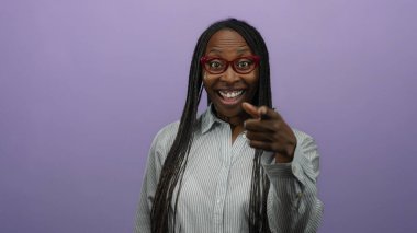 Kıvırcık saçlı, soyutlanmış mor arka planda gülümseyen gözlüklü ve çizgili tişörtlü bir kadın neşe ve güven ifade ediyor..