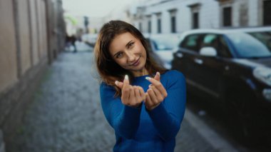 Mavi kazaklı genç bir kadın park etmiş arabalarla şehir caddesinde parmakları olan kalp sembolleri yapıyor..