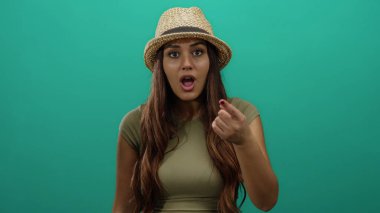 Şapka takan İspanyol kadın şaşkın bir ifadeyle izole edilmiş yeşil arka planı işaret ederek canlı bir zemin üzerinde şaşırtıcı ve meraklı bir şekilde ifade ediyor..