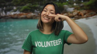 Deniz kenarında gülümseyen genç Çinli bir kadın, gönüllü bir tişört giyiyor, burnunu işaret ediyor, açık mavi suyla sakin bir kumsalda gönüllü olma ruhunu somutlaştırıyor..
