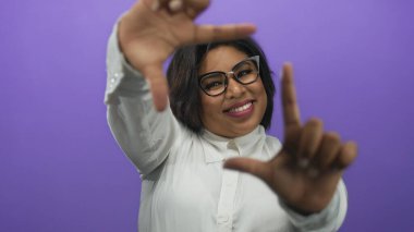 Elleriyle gülümseyen ve çerçeveleyen bir kadın, mor arka planda gözlük takan, izole bir ortamda pozitiflik ve yaratıcılık taşıyan bir kadın..