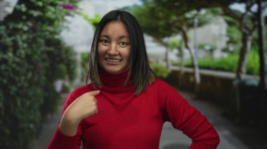 Parkta gülümseyen kırmızı kazaklı genç Çinli kadın kendini ağaçlar ve çalılarla göstererek açık havada hareketli bir ortam yaratıyor..