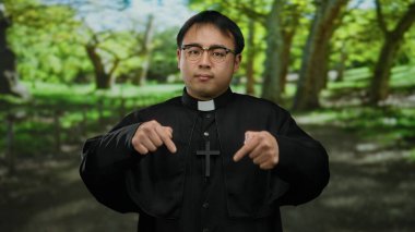 Güneşli bir parkta arka planda ağaçlar olan gözlüklü bir rahip, sakin bir açık hava manzarası sergiliyor..