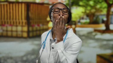 Beyaz önlüklü ve steteskoplu Afrikalı Amerikalı bir kadın güneşli bir parkta öpücükler yolluyor..