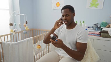 Yatak odasındaki genç adam, evde telefonla şişeyi tutuyor. Bebek beşiğinin yanında, etrafı battaniye ve dekorla çevrili..