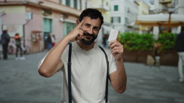 Sakallı, elinde kredi kartı olan İspanyol bir adam şehir sokaklarında gülümsüyor..