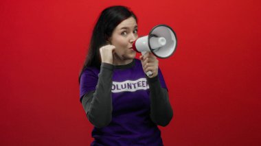 Kadın megafonla tutkuyla bağırıyor. Gönüllü gömleği ile kırmızı arka plana vuruyor. Enerjik bir gösteride vurgu yapmak için yumruğunu sıkıyor..