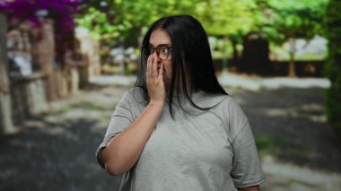 Gri gömlekli, kıvrımlı genç bir kadın güneşli parkta duruyor. Etrafı canlı ağaçlarla çevrili, arka planı bulanık, dış görünüşü ve kendine güveni somutlaştırıyor..