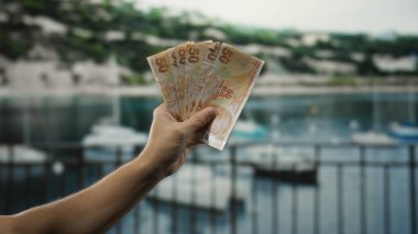 Deniz kıyısında elinde Türk lirası banknotları olan bir adam arka planda tekneler, deniz kenarında açık hava finansal faaliyetlerinin özünü yakalıyor..