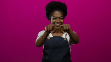 Önlüklü genç kadın gülümsüyor ve doğrudan kameraya bakıyor. İzole edilmiş pembe arka planda, kendine güven ve yakınlık yayıyor.