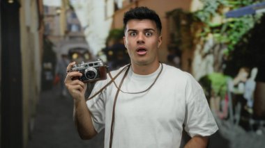 Arka planda bulanık bir kamera tutan genç adam gündüz vakti açık havada beyaz gömleğiyle oyun oynuyor..