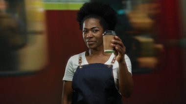 Tren istasyonunda kahve fincanı tutan, geçmişi bulanık, gülümseyen ve yoğun bir seyahat ortamında misafirperverliği temsil eden önlüklü bir kadın..