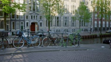 Arka planda Amsterdam 'ın ikonik kanal evleri olan kaldırım taşı caddesi boyunca park etmiş bulanık bisikletlerle sakin, bokeh dolu bir şehir manzarası yakalıyorlar..