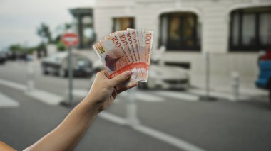 Elde tutulan Norveç kronu, hareketli bir şehir caddesinde renkli banknotlar sergiliyor, açık şehir finans veya ekonomik bağlamı vurguluyor.