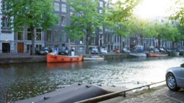 Hollanda kıyısı boyunca park halindeki araçlar ve teknelerin bulunduğu bulanık kanallar, güneşli bir ortam içinde tipik bir sokak manzarası ve mimari unsurlar sergiliyor..