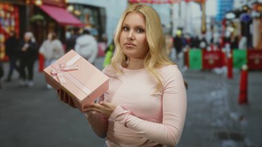 Şehrin hareketli sokak arkaplanında pembe hediye kutusu tutan kadın, şehrin canlı atmosferindeki günümüze dair karışık duygular paylaşıyor..