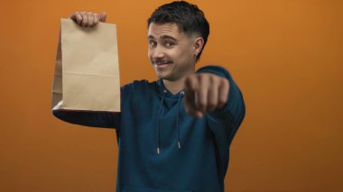 İzole edilmiş turuncu arka planda kameraya işaret eden İspanyol genç adam nişan ve etkileşim öneriyor.